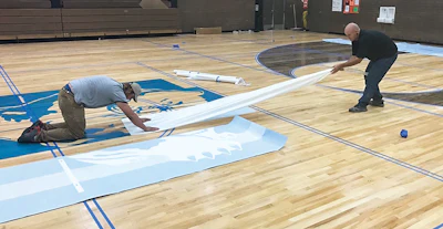 An important part of gym finish work is understanding how to use stencils. Here we’re applying stencils prior to painting a center logo.