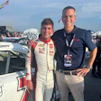 Race car driver in white suit standing next to team member at motorsport event