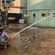 Firefighters spraying water from a hose near industrial storage tanks