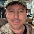 Person in olive green cap and beige jacket smiling in an indoor workspace