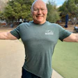 Smiling bald man in gray Rubio Monocoat t-shirt standing outdoors with trees behind