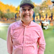 Smiling man in pink shirt and green cap standing outdoors with autumn trees