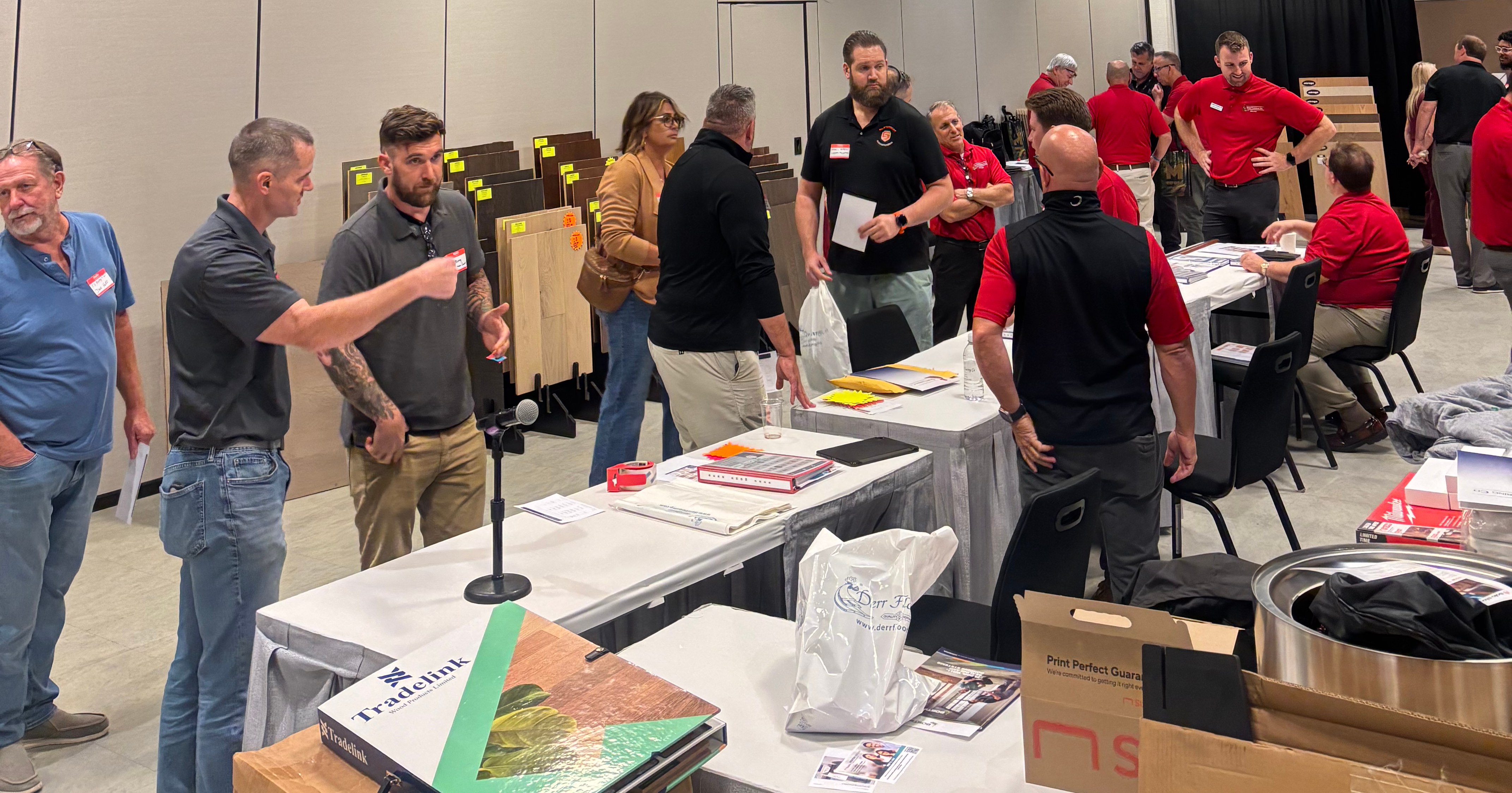 Group of people networking at an indoor event with tables displaying materials and signage in a convention center.