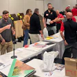 Group of people networking at an indoor event with tables displaying materials and signage in a convention center.