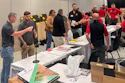 Group of people networking at an indoor event with tables displaying materials and signage in a convention center.