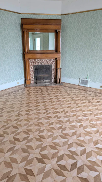 Empty living room with wooden fireplace mantel, mirror, decorative brick surround, patterned wood floor, and light blue wallpaper.