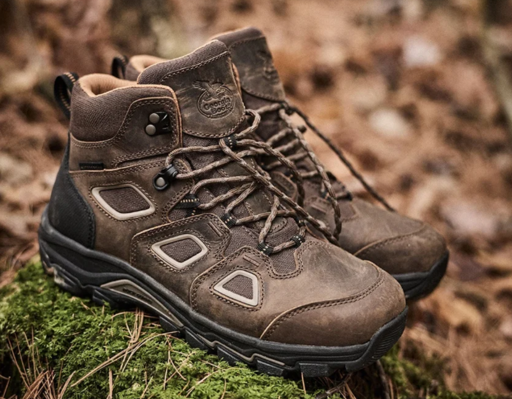 Pair of brown suede hiking boots with black soles positioned on moss and rocky terrain