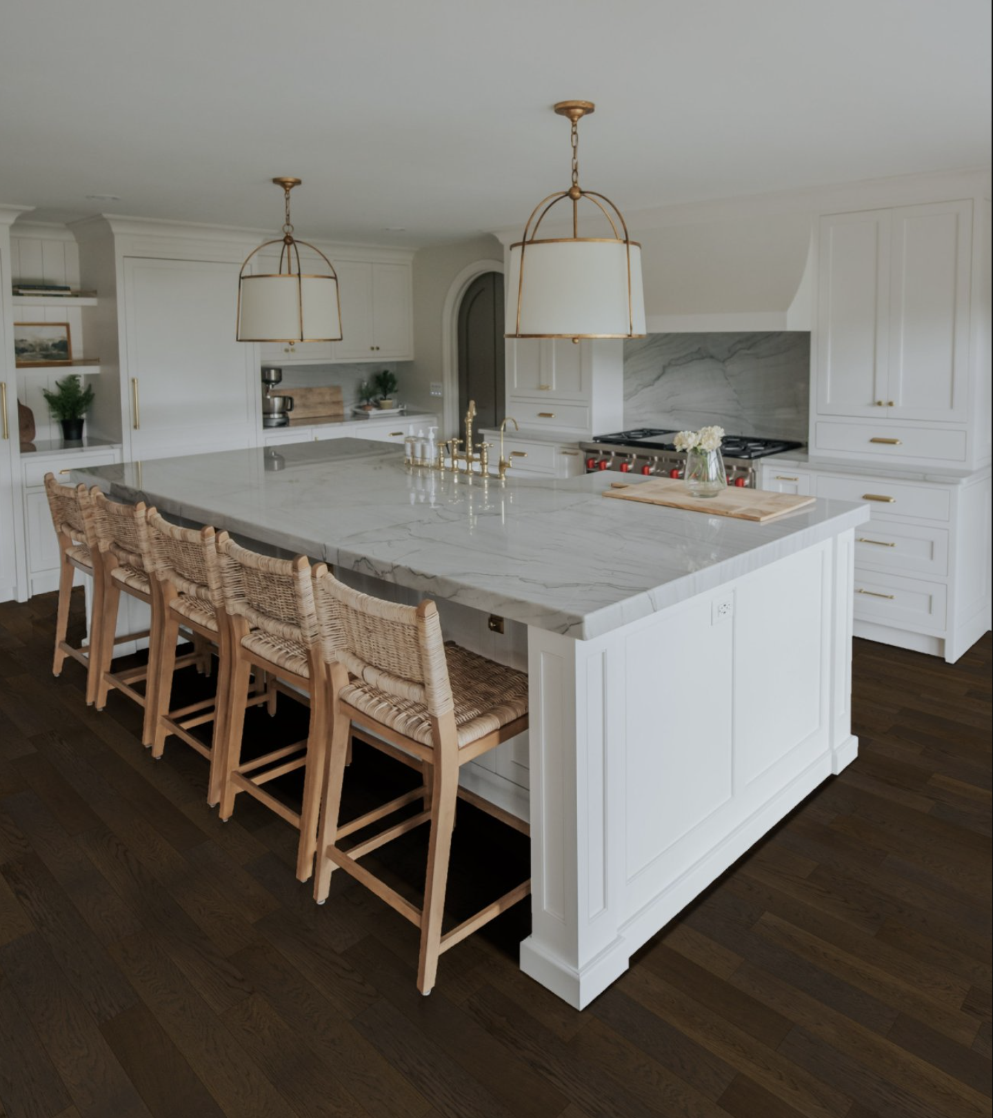 Modern white kitchen with marble island, woven bar stools, brass pendant lights, and dark wood flooring
