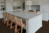 Modern white kitchen with marble island, woven bar stools, brass pendant lights, and dark wood flooring