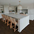 Modern white kitchen with marble island, woven bar stools, brass pendant lights, and dark wood flooring