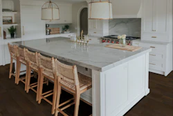 Modern white kitchen with marble island, woven bar stools, brass pendant lights, and dark wood flooring