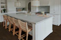 Modern white kitchen with marble island, woven bar stools, brass pendant lights, and dark wood flooring
