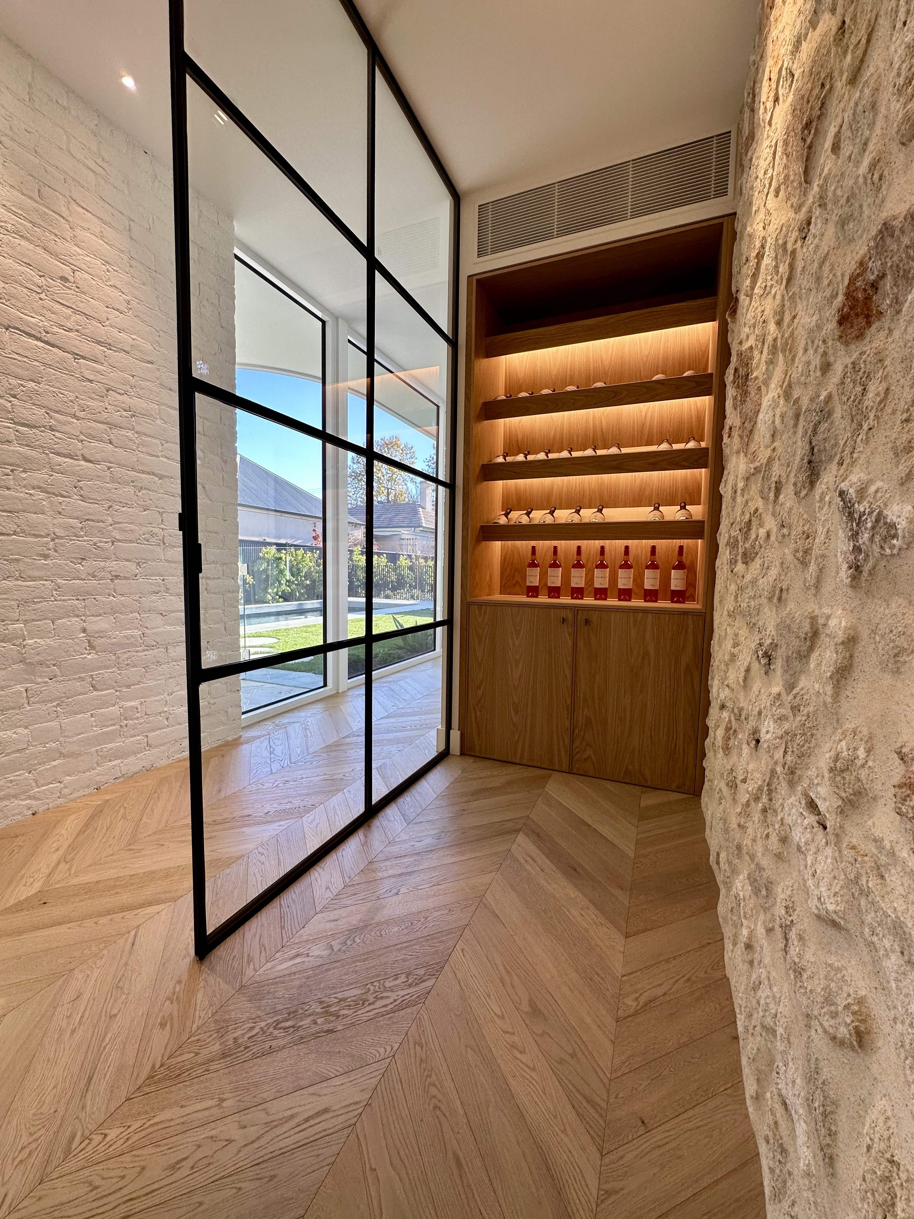 Modern entryway with black-framed glass doors, white brick walls, chevron flooring, and illuminated wooden wine display cabinet