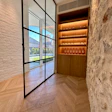 Modern entryway with black-framed glass doors, white brick walls, chevron flooring, and illuminated wooden wine display cabinet