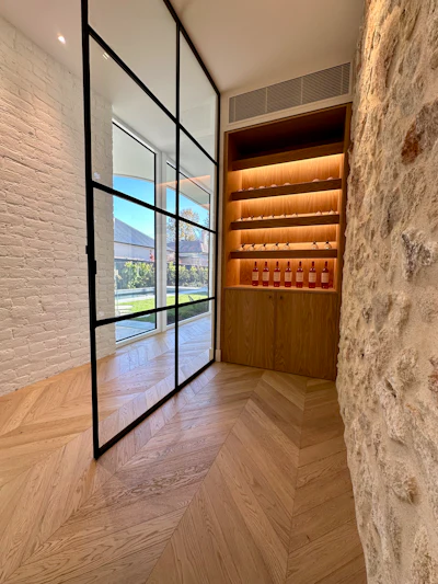 Modern entryway with black-framed glass doors, white brick walls, chevron flooring, and illuminated wooden wine display cabinet