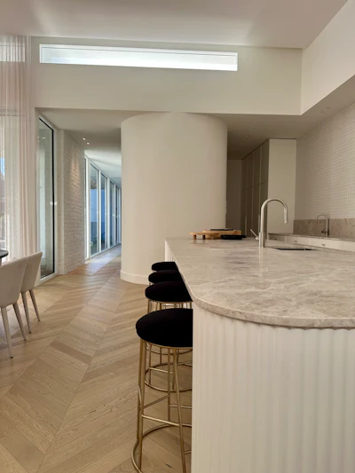 Modern kitchen with curved marble island, black bar stools, chevron flooring, and natural light from skylight window