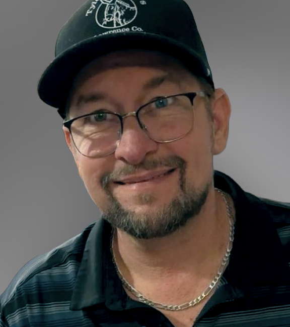 Man wearing glasses and black baseball cap with logo, smiling at camera against gray background