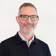 Professional headshot of middle-aged man wearing glasses and dark jacket with checkered shirt