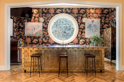 Luxury bar interior with brass studded counter, circular mirror, and patterned wallpaper backdrop
