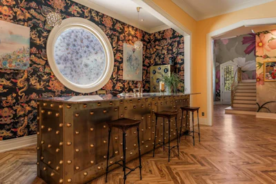 Modern bar interior with studded bronze counter, round mirror, dark floral wallpaper, and yellow accent walls