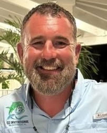 Smiling man with beard wearing light blue shirt with logo, indoor setting with plants Headshot