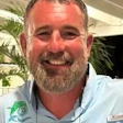 Smiling man with beard wearing light blue shirt with logo, indoor setting with plants